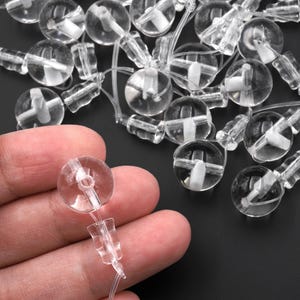 Peut inclure: Un ensemble de tendeurs de cordon en plastique transparent pour stores, chacun doté d'un composant supérieur sphérique avec un trou central et d'une base rectangulaire connectée, conçus pour fixer et ajuster les cordons de tirage fins.