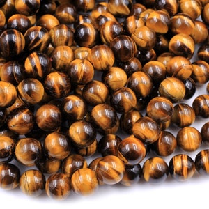 May include: A close-up of a pile of round, polished tiger's eye beads. The beads are a mix of golden brown and dark brown colors with a shimmering, chatoyant effect.