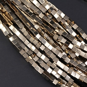 May include: Close-up of a gold-colored necklace or bracelet. The jewelry is made of small, square, metallic beads strung together. The beads have a reflective surface, and the overall effect is shiny and elegant. The background is a dark, solid color.