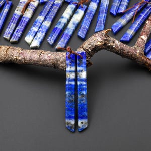 May include: Rectangular lapis lazuli gemstone beads with a deep blue color, white veining, and flecks of gold. The beads are strung on brown cord and displayed on a tree branch against a dark background. These beads are ideal for jewelry making.