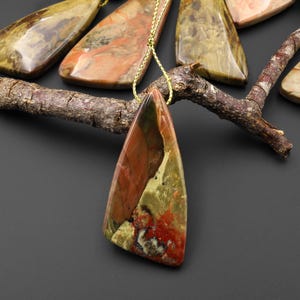 May include: A selection of gemstone pendants with a triangular form and a gold chain. The pendants display a mix of colours, including red, green, brown, and cream. The pendants are presented on a dark surface with a wooden branch.