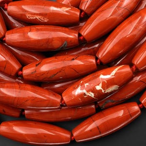 May include: Close-up of a collection of polished, elongated red jasper beads. Each bead is barrel-shaped with natural veining and patterns in darker shades. The beads are strung together with red thread, creating a rich, textured appearance, ideal for jewelry making.