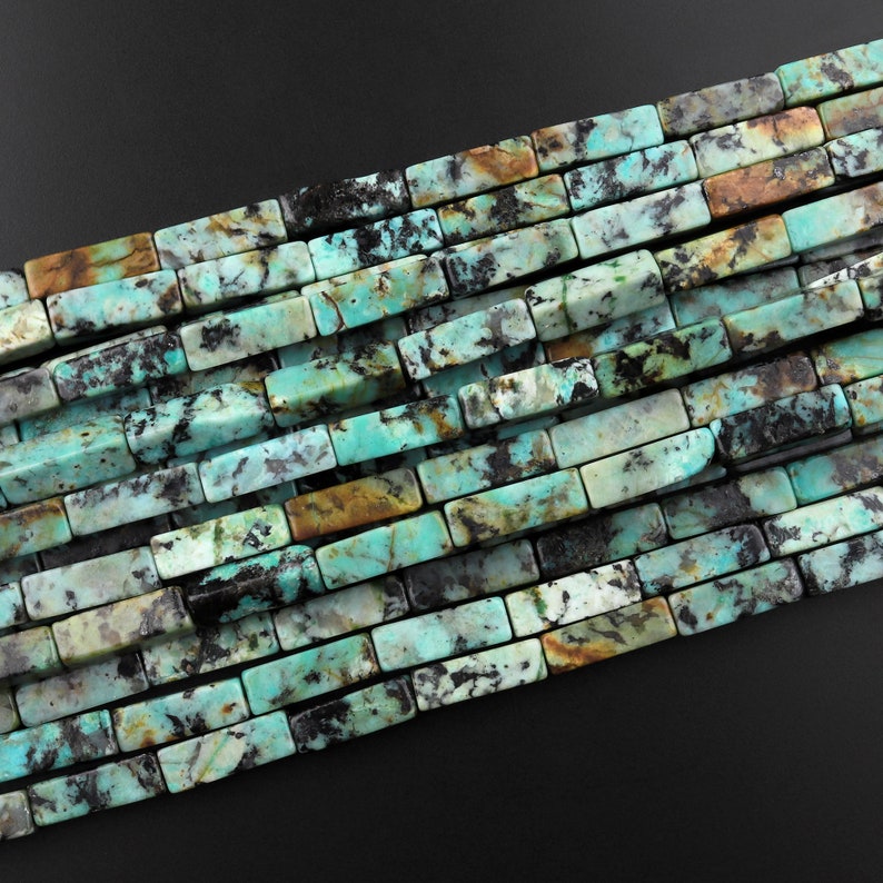 May include: A strand of turquoise gemstone beads, each bead is a rectangular shape with a mottled pattern of blue, green, brown, and black.