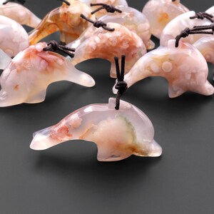 May include: A group of pink and white agate dolphin pendants with brown cords. The dolphins are carved from natural agate and have a variety of patterns and colors.