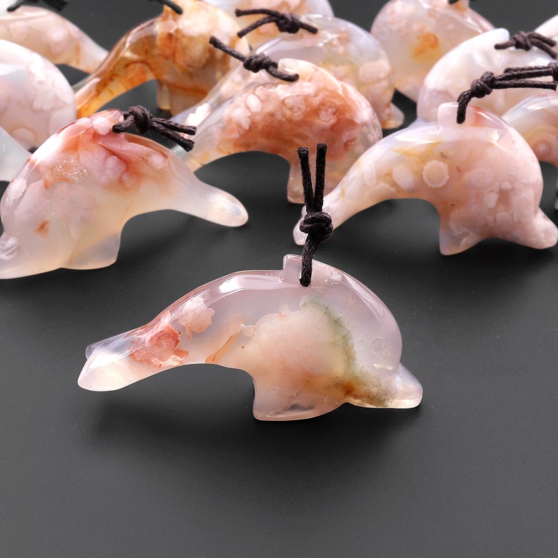 May include: A group of pink and white agate dolphin pendants with brown cords. The dolphins are carved from natural agate and have a variety of patterns and colors.