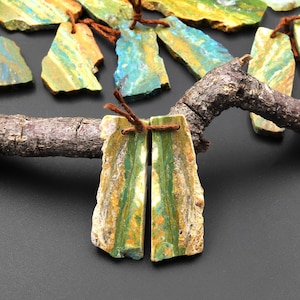 May include: A pair of green and brown stone earrings with a natural, irregular shape. The earrings are attached to brown leather cords.