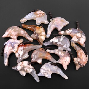May include: A collection of 12 agate dolphin pendants with white and pink agate patterns. The pendants are carved in the shape of dolphins and have a brown cord attached.