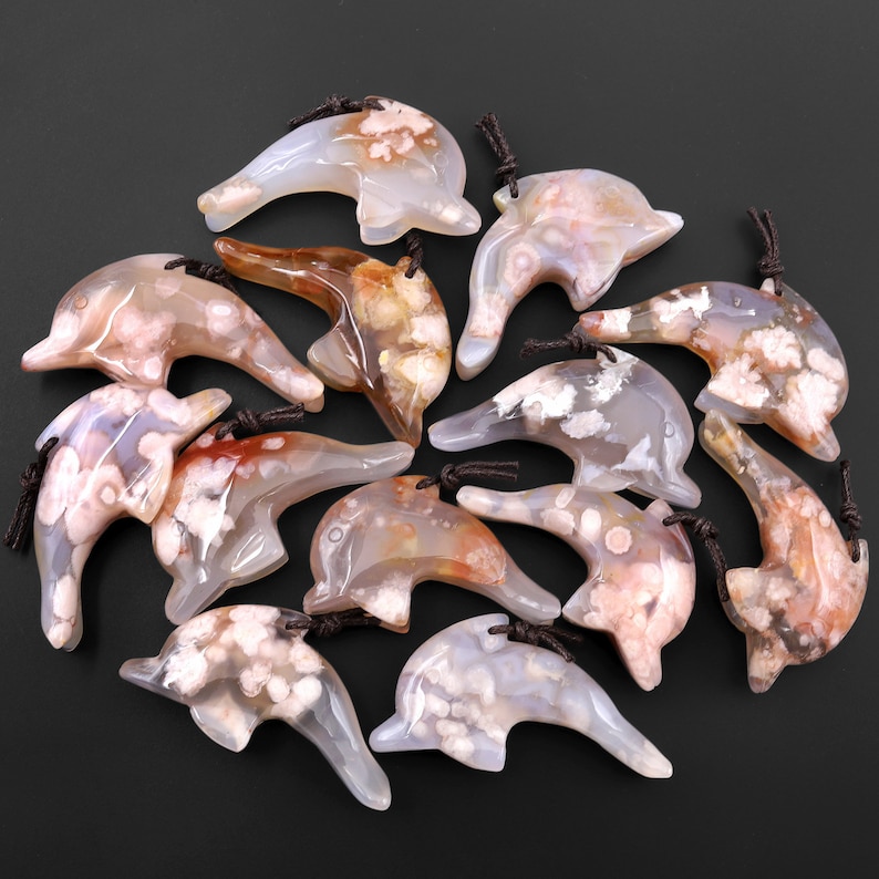 May include: A collection of 12 agate dolphin pendants with white and pink agate patterns. The pendants are carved in the shape of dolphins and have a brown cord attached.