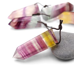 Puede incluir: Un primer plano de un colgante de cristal de fluorita multicolor con un cordón de cuero marrón. El cristal es de color morado oscuro, rosa y amarillo con rayas blancas. El colgante está apoyado sobre una piedra gris.