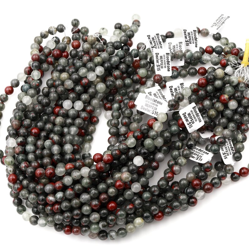 May include: A close-up of a pile of round, green, red, and white bloodstone beads. The beads are all about 8mm in diameter and are strung on a clear string. The beads are labeled with the text "Bloodstone MSRP$30 Dealer:$15".