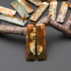 May include: Rectangular gemstone pendants in shades of brown, green, and cream, with natural patterns. Each pendant is approximately 2 inches long and has a small hole for stringing. The pendants are displayed on a dark surface.