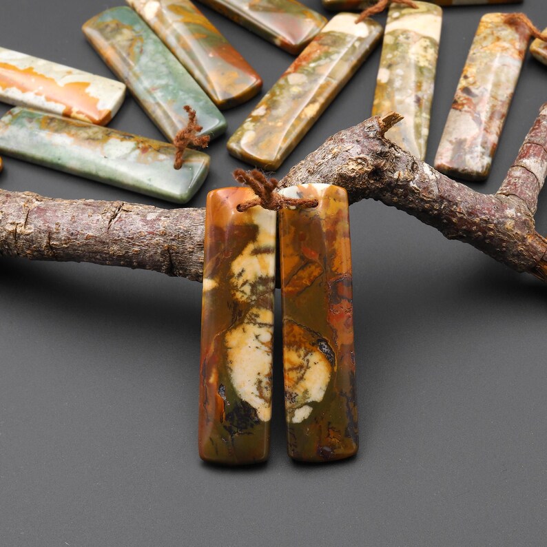 May include: Rectangular gemstone pendants in shades of brown, green, and cream, with natural patterns. Each pendant is approximately 2 inches long and has a small hole for stringing. The pendants are displayed on a dark surface.