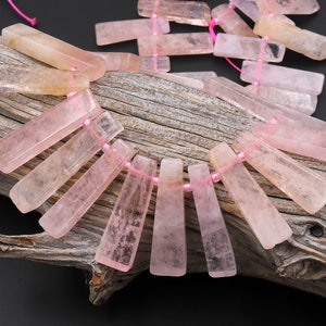 May include: Rose quartz gemstone beads in a rectangular shape, strung on pink thread. The beads are a pale pink color and are displayed on a piece of weathered wood. The beads are arranged in a fan-like pattern.