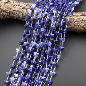 Op de afbeelding: Close-up van meerdere strengen blauwe, vlindervormige kralen. De kralen zijn geregen op zilverkleurig draad. De kralen zijn diepblauw met lichtere blauwe en witte accenten. Op de achtergrond een stuk hout.