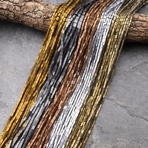 May include: An assortment of metallic beads in gold, silver, bronze, and black. The small, square-shaped beads are strung together, creating a textured display. The beads are arranged in parallel lines. The image shows a variety of colours.