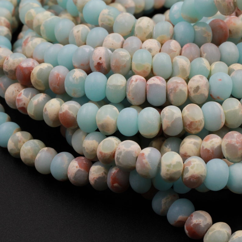 May include: Strands of gemstone beads in shades of pale blue, cream, and reddish-brown. The beads are oval-shaped and have a matte finish. The beads are strung together, creating a textured and colorful pattern. Ideal for jewelry making.