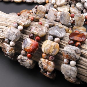 May include: Close-up of several beaded bracelets featuring large, irregularly shaped stones in shades of gray, brown, and red. The bracelets are strung with smaller, round beads in complementary colors. The stones have unique patterns.