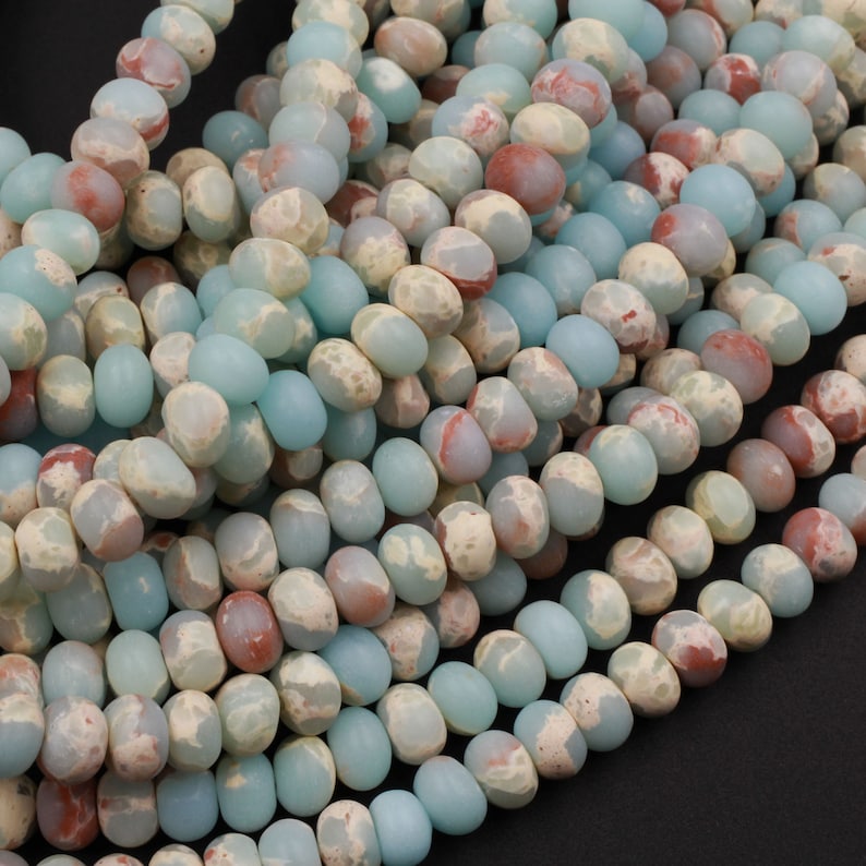 May include: Close-up of strands of gemstone beads. The beads are a mix of pale blue, cream, and reddish-brown colors. Each bead is oval-shaped and has a slightly irregular surface. The beads are strung together, creating a textured and colorful display.