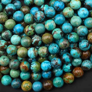 May include: A collection of round turquoise beads featuring natural variations in color, ranging from bright sky blue to deep teal and earthy brown matrix patterns. Each bead has a smooth, polished surface with unique veining throughout the cluster.
