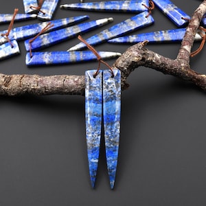 May include: Blue lapis lazuli gemstone pendants with a rectangular shape and pointed ends. The pendants are strung with brown cord. Other lapis lazuli pieces are scattered in the background. The gemstones have a mix of blue and gray tones.