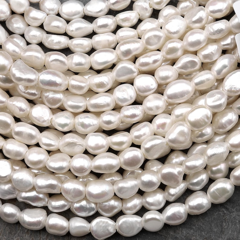 Puede incluir: Primer plano de una colecci&oacute;n de perlas de agua dulce blancas cremosas de forma irregular. Las perlas est&aacute;n ensartadas, creando una superficie texturizada y luminosa. Las perlas tienen un aspecto natural y org&aacute;nico.