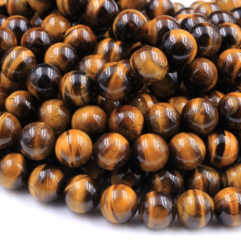 May include: A close-up of a pile of round, polished tiger's eye beads. The beads are a mix of golden brown and black colors with a shimmering, iridescent effect.