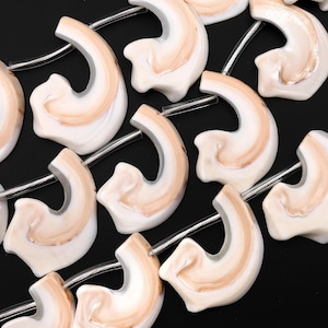 Puede incluir: Una hebra de cuentas de cerámica blancas y beige con un patrón de remolino. Las cuentas tienen forma de oreja curva y están enhebradas en un alambre plateado.