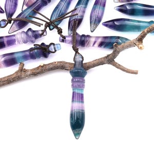 May include: A collection of colorful fluorite pendulums with a variety of purple, blue, and green hues. Each pendulum is suspended by a brown cord. The pendulums are arranged on a white background, some resting on a small branch.