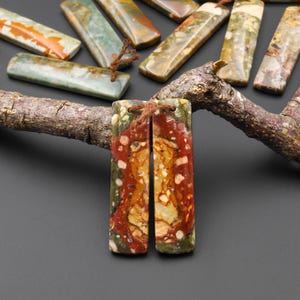 May include: Rectangular gemstone pendants with a mix of red, green, and yellow hues. The pendants are strung with brown cord and are displayed on a dark surface. Other similar stones are visible in the background.
