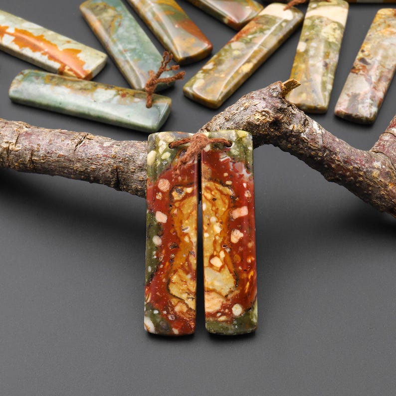 May include: Rectangular gemstone pendants with a mix of red, green, and yellow hues. The pendants are strung with brown cord and are displayed on a dark surface. Other similar stones are visible in the background.