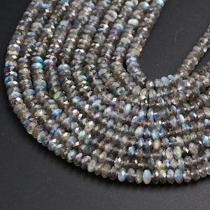 May include: A strand of faceted labradorite beads, with a dark gray color and iridescent blue and green flashes. The beads are round and have a smooth, polished finish.