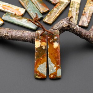 May include: Two rectangular, polished stone pendants with a brown cord, featuring a mix of orange, brown, white, and green hues. Other similar stone pieces are arranged on a wooden branch, showcasing natural variations in color and pattern.