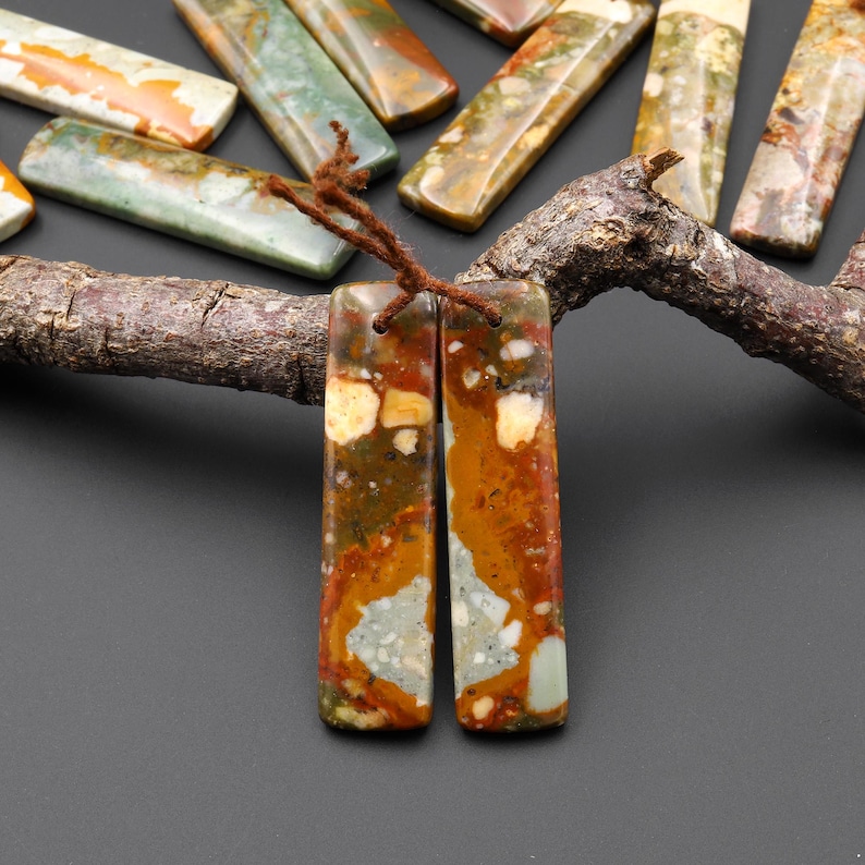 May include: Two rectangular, polished stone pendants with a brown cord, featuring a mix of orange, brown, white, and green hues. Other similar stone pieces are arranged on a wooden branch, showcasing natural variations in color and pattern.