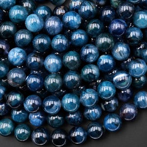 May include: A close-up shot of numerous blue-green gemstone beads. The beads are round and polished, with a glossy finish. The color varies from deep teal to lighter shades of blue, creating a gradient effect. These beads are suitable for jewelry making.