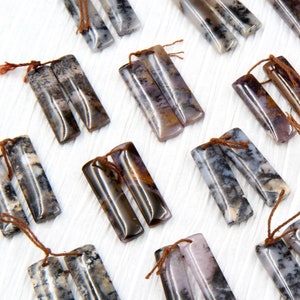 May include: A set of 10 pairs of agate gemstone pendants, each pair featuring two rectangular pieces with a brown, black, and white marbled pattern. The pendants have a natural, polished finish and are strung with brown cords.