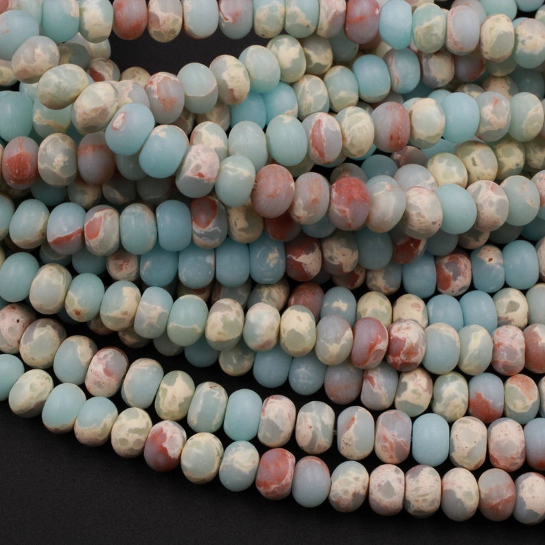 May include: Close-up of strands of gemstone beads in shades of blue, cream, and reddish-brown. The beads are oval-shaped and have a matte finish. The beads are strung together, creating a textured and colorful pattern.