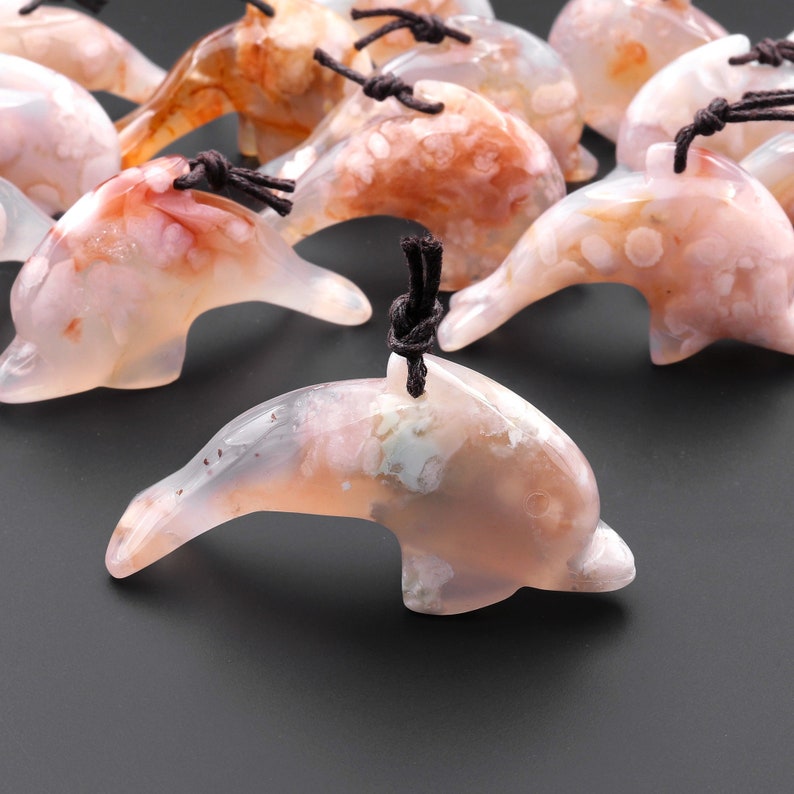 May include: A group of pink and white agate dolphin pendants with brown cords. The dolphins are carved from natural agate and have a unique, marbled pattern.