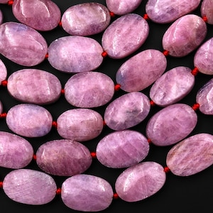 May include: Close-up of a strand of oval-shaped, light purple gemstones. The stones are strung on a red cord, creating a necklace or bracelet. The gemstones have a polished, smooth surface.