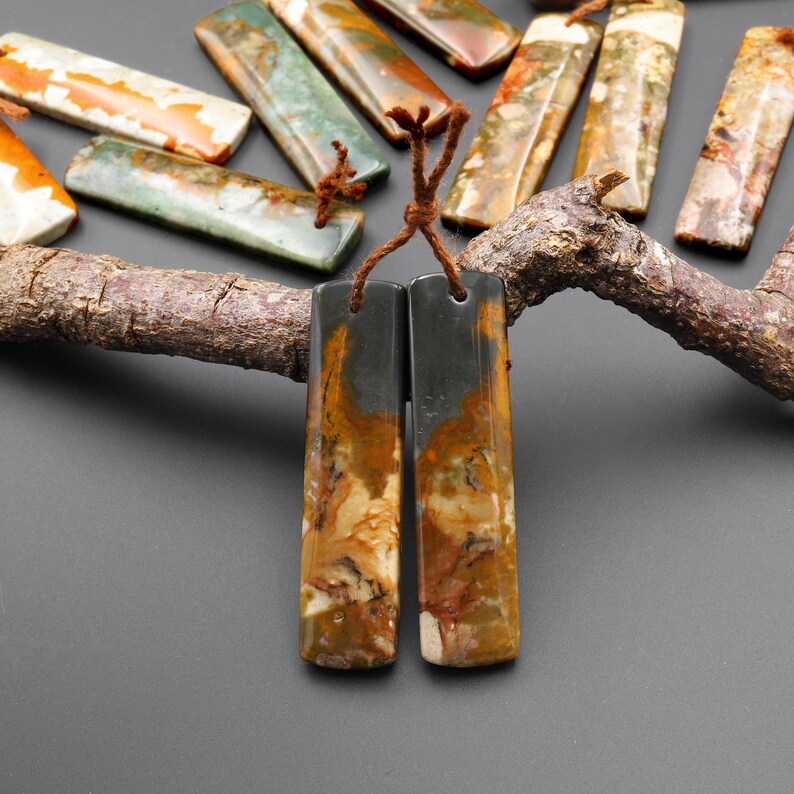 May include: A collection of rectangular gemstone pendants in shades of brown, orange, green, and gray. Two pendants are in the foreground, connected by a brown cord. The pendants are displayed on a dark gray surface with a wooden branch.