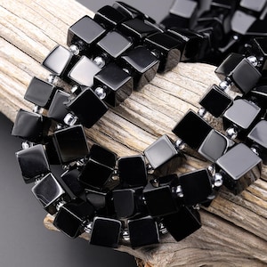 May include: A close-up of a black necklace with square beads. The beads are strung together with small silver-colored beads. The necklace is displayed on a piece of wood.