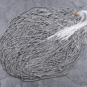 May include: A strand of silver-colored beads, likely made of hematite, arranged in a circular pattern on a gray surface. The beads are small and uniform in size, creating a shimmering effect.