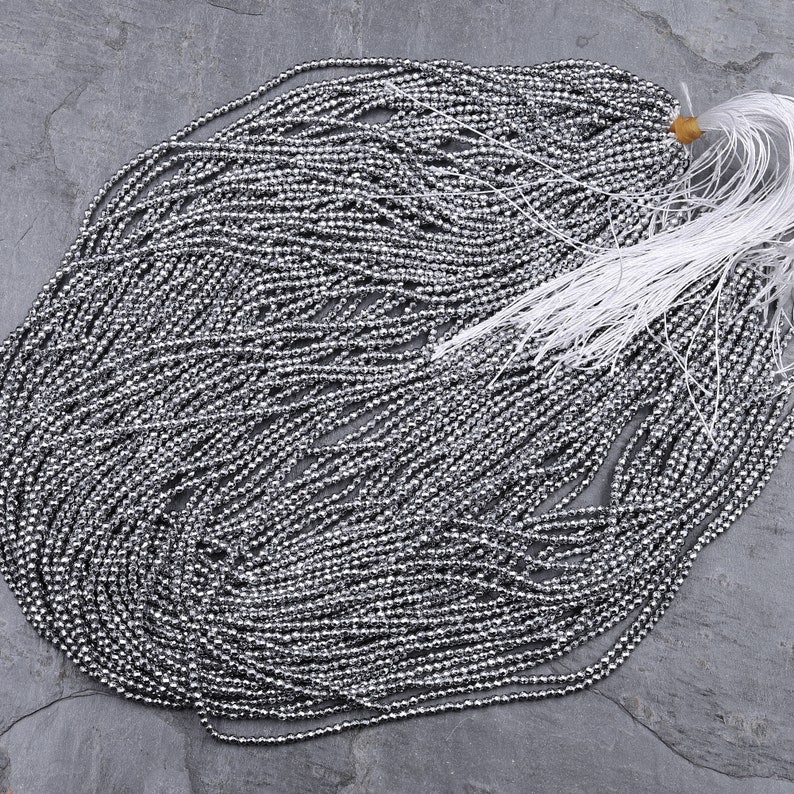 May include: A strand of silver-colored beads, likely made of hematite, arranged in a circular pattern on a gray surface. The beads are small and uniform in size, creating a shimmering effect.
