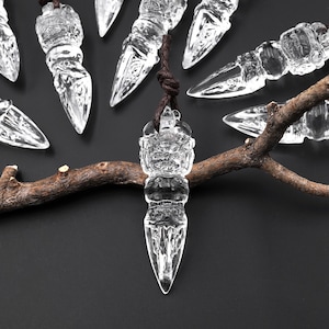 May include: Clear crystal pendants with a dagger-like design, suspended from brown cords. The pendants are arranged on a dark surface, with a brown branch adding a natural element. The crystal pendants have intricate details and a sharp point.