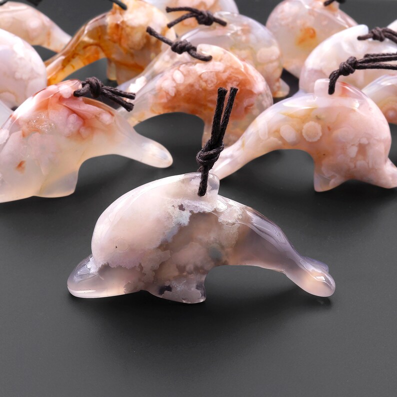 May include: A group of white agate dolphin pendants with brown cords. The dolphins have a mottled, cloudy appearance with white, gray, and pink hues.