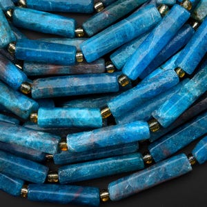 May include: A collection of cylindrical blue apatite gemstone beads arranged in parallel rows. Each bead features a natural, semi-translucent texture with deep azure and teal hues, separated by small, reflective gold-toned metallic spacer beads.