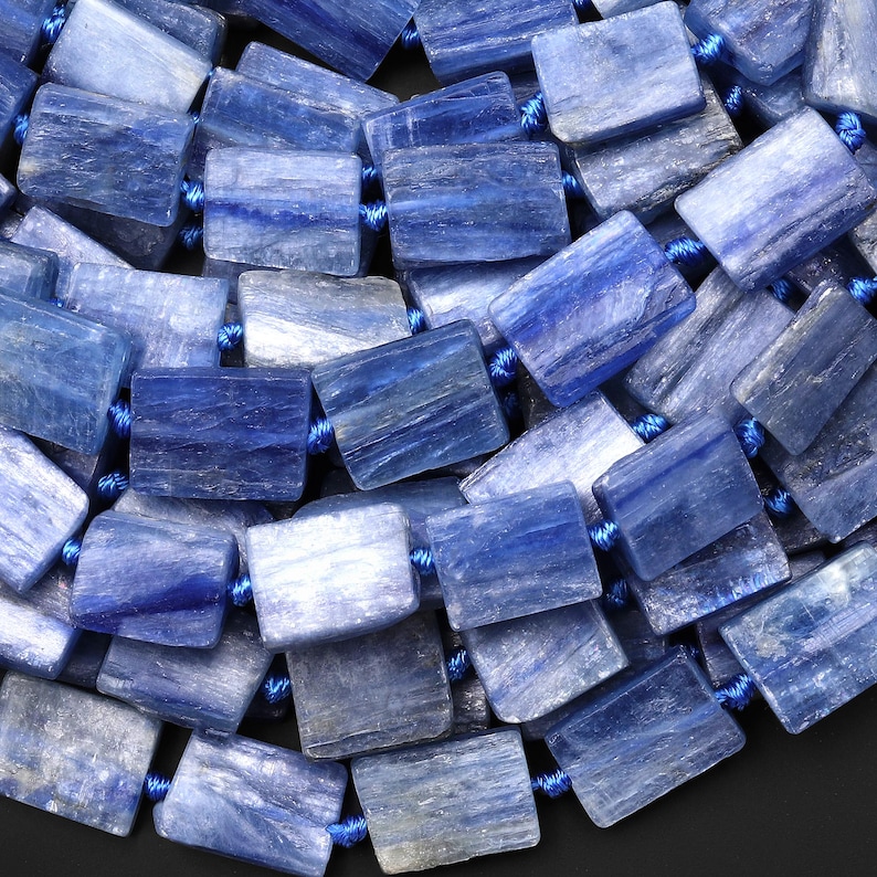 Puede incluir: Primer plano de un collar hecho de cuentas rectangulares de kianita azul. Las cuentas var&iacute;an en tonos de azul, de claro a &iacute;ndigo profundo, y est&aacute;n ensartadas en un cord&oacute;n azul. La piedra natural tiene una apariencia texturizada.