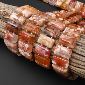 May include: A multi-strand bracelet composed of rectangular, polished agate beads in shades of orange, cream, and brown. The beads feature natural marbled patterns and are connected by small silver-toned metal spacers, creating a layered appearance.