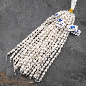 Puede incluir: Hilos de perlas blancas de forma irregular, probablemente de agua dulce, est&aacute;n agrupados. Cada hilo est&aacute; ensartado con un hilo visible. Peque&ntilde;as etiquetas blancas con el texto "INTRINSIC TRADING" est&aacute;n adjuntas.