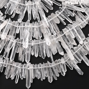 May include: Close-up of multiple strands of clear quartz crystal points, strung together on white thread. The crystals are translucent and vary in size and shape, creating a natural, textured appearance. The background is black, highlighting the crystals.