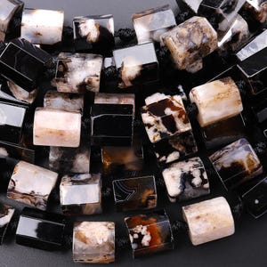 May include: Close-up of a collection of polished, rectangular beads. The beads are a mix of black, white, and brown, with unique patterns and textures. They are strung together on a black cord, suggesting a necklace or bracelet.